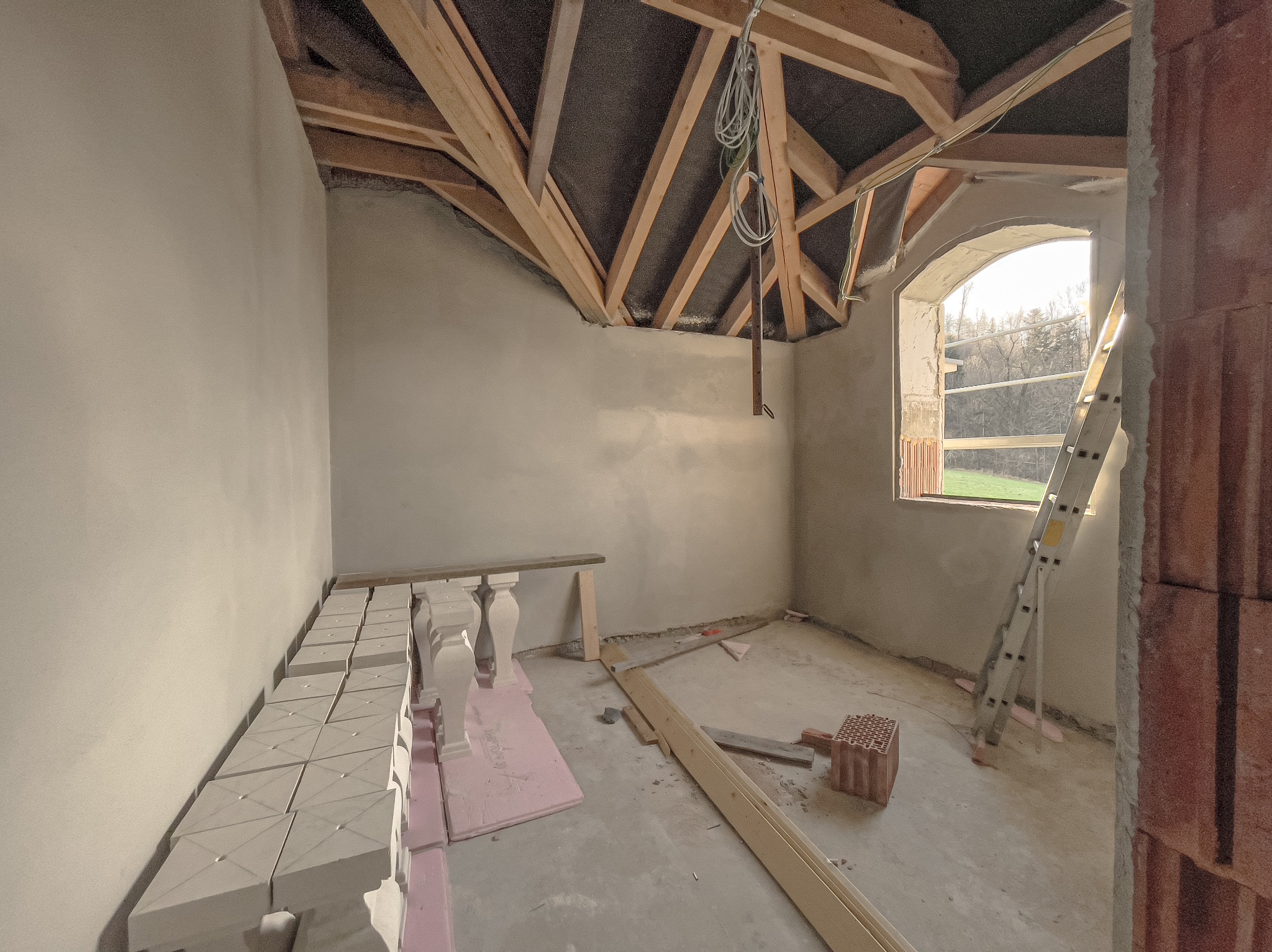 Wnętrze w trakcie prac wykończeniowych: ściany po nałożeniu tynku, surowy drewniany strop, okno z łukowym nadprożem i drabina oparta o mur, fragment balustrady na podłodze.