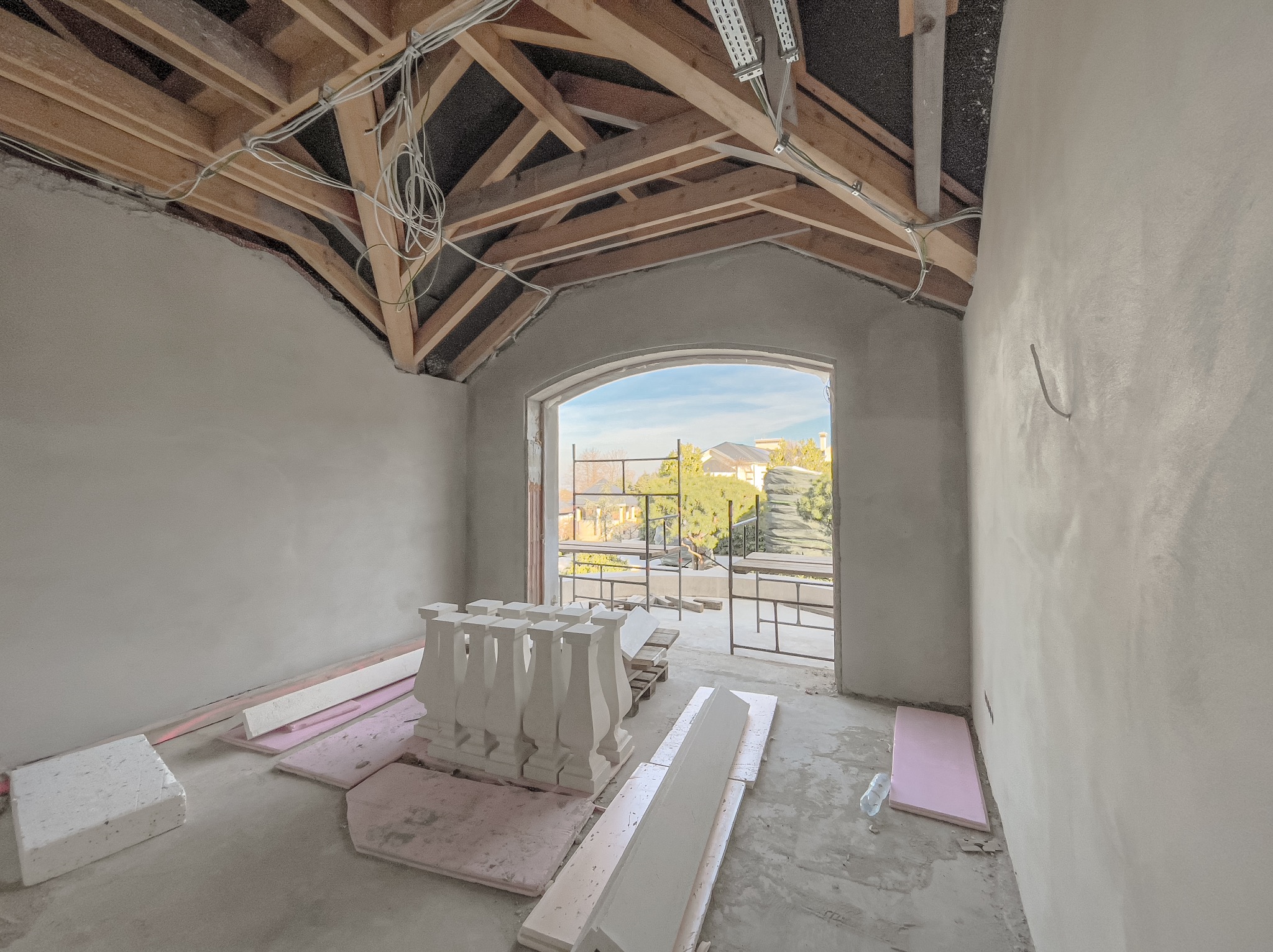 Wnętrze w trakcie tynkowania maszynowego z widoczną konstrukcją drewnianą dachu, przewodami elektrycznymi oraz elementami balustrady balkonowej na podłodze, widok na zewnątrz przez otwór okienny.