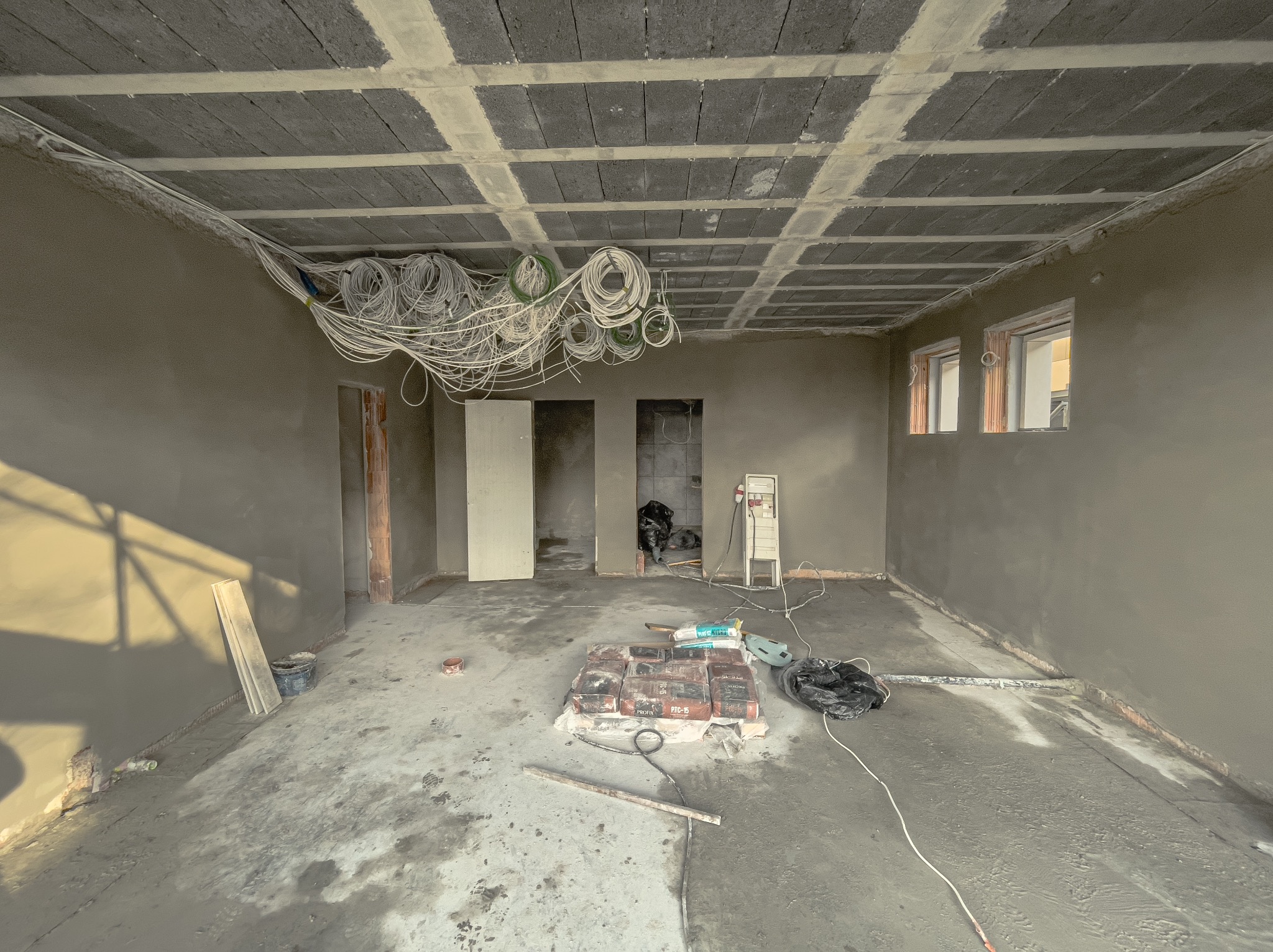 Wnętrze w trakcie prac wykończeniowych: świeżo nałożone szare tynki na ścianach, zwisające kable elektryczne z sufitu z betonowych bloków, worki z zaprawą na podłodze, widoczne dwa okna...