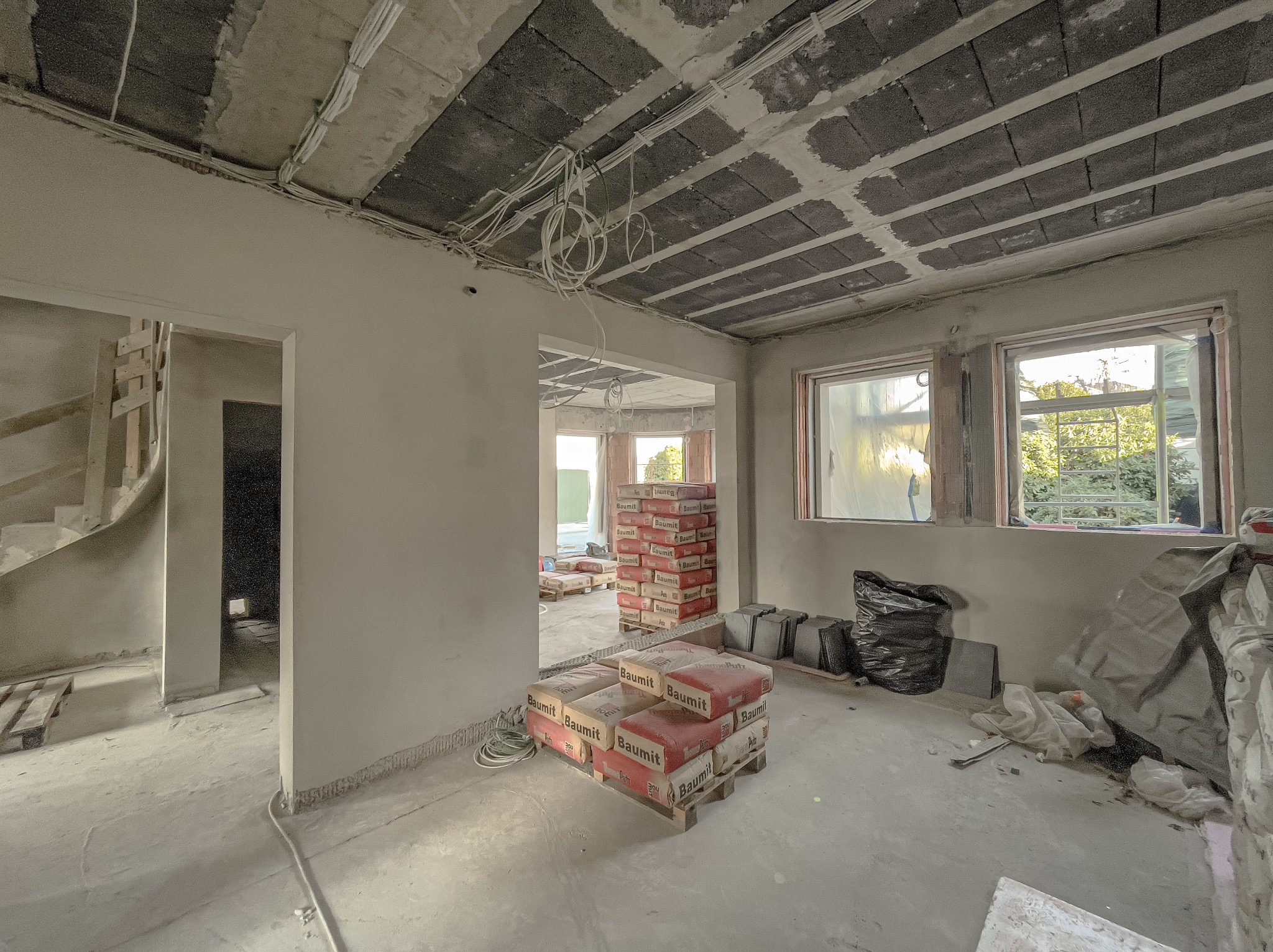 Wnętrze w stanie surowym z widocznymi workami zaprawy Baumit na paletach, instalacją elektryczną pod sufitem i oknami zabezpieczonymi folią.