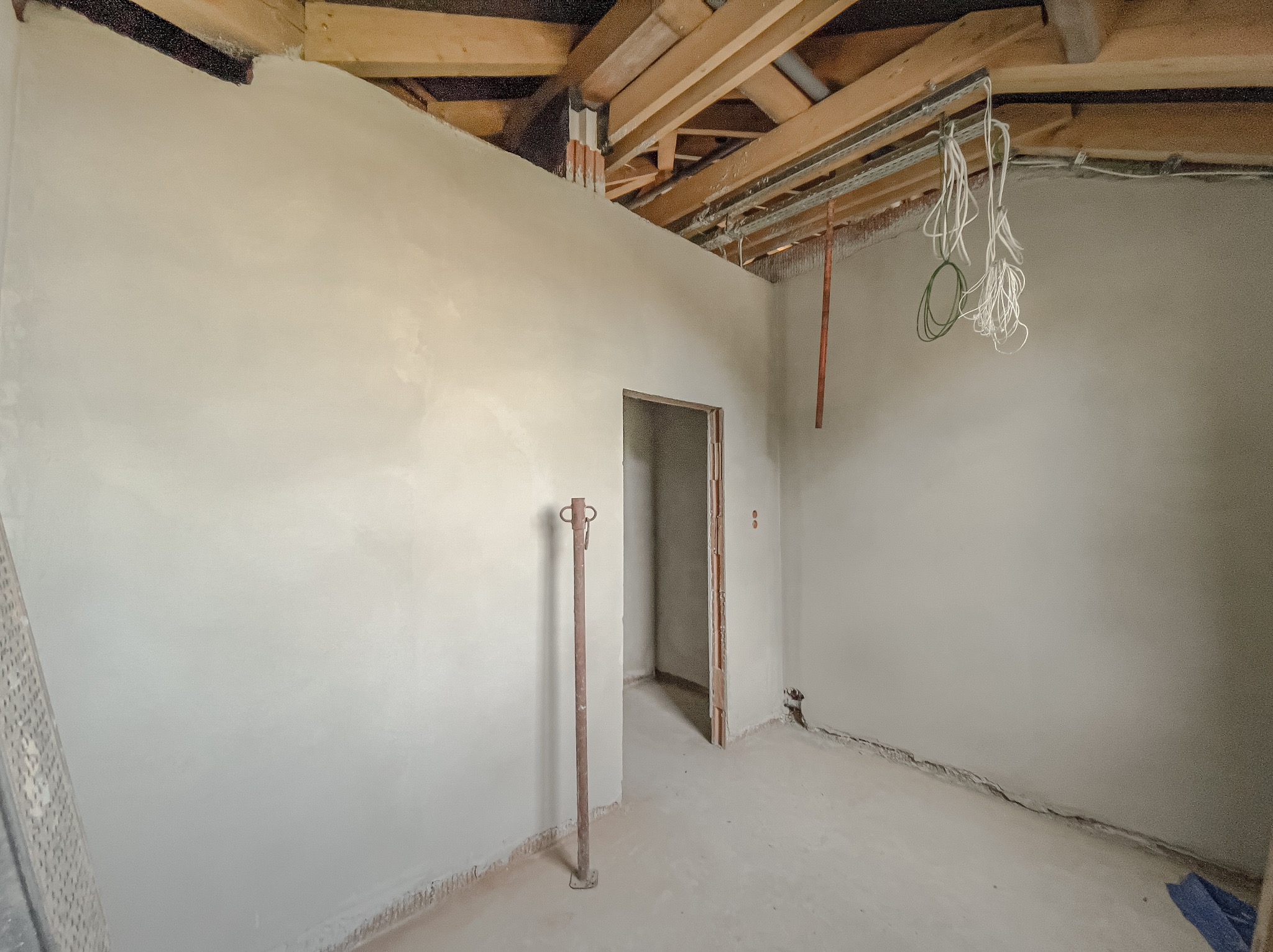 Świeżo otynkowane maszynowo ściany w pomieszczeniu z widocznym drewnianym stropem, wystającymi przewodami i metalową podporą.