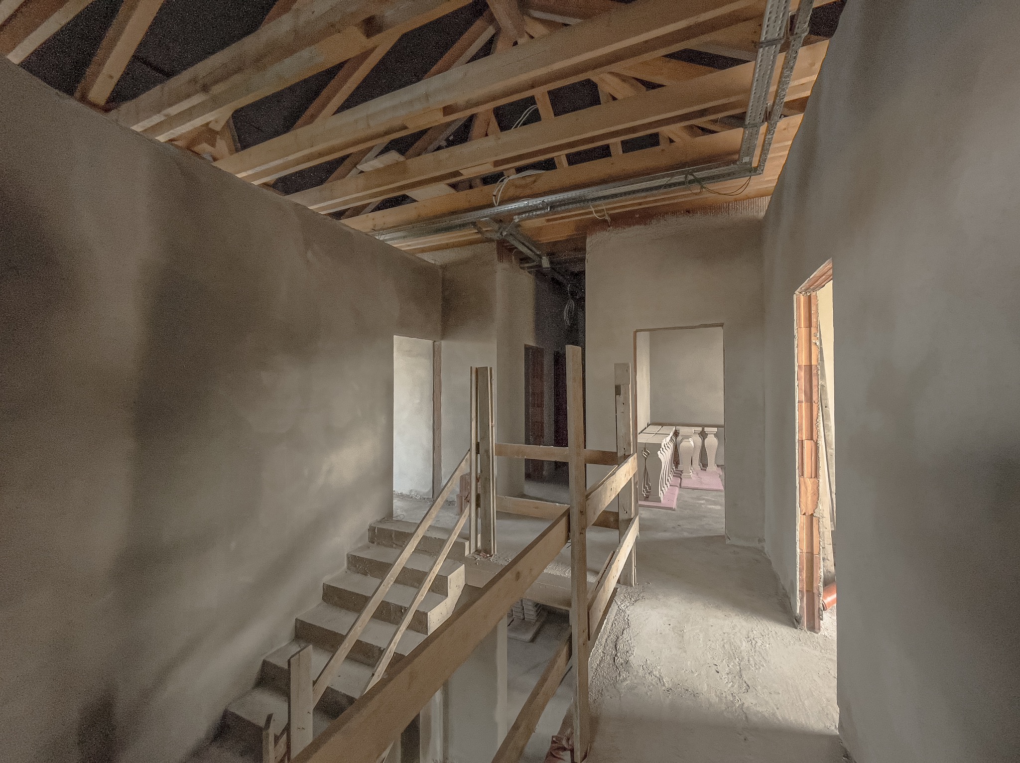 Wnętrze budynku w trakcie prac wykończeniowych, ściany pokryte świeżym tynkiem, widoczne drewniane elementy konstrukcji dachu oraz schody z drewnianą barierką.