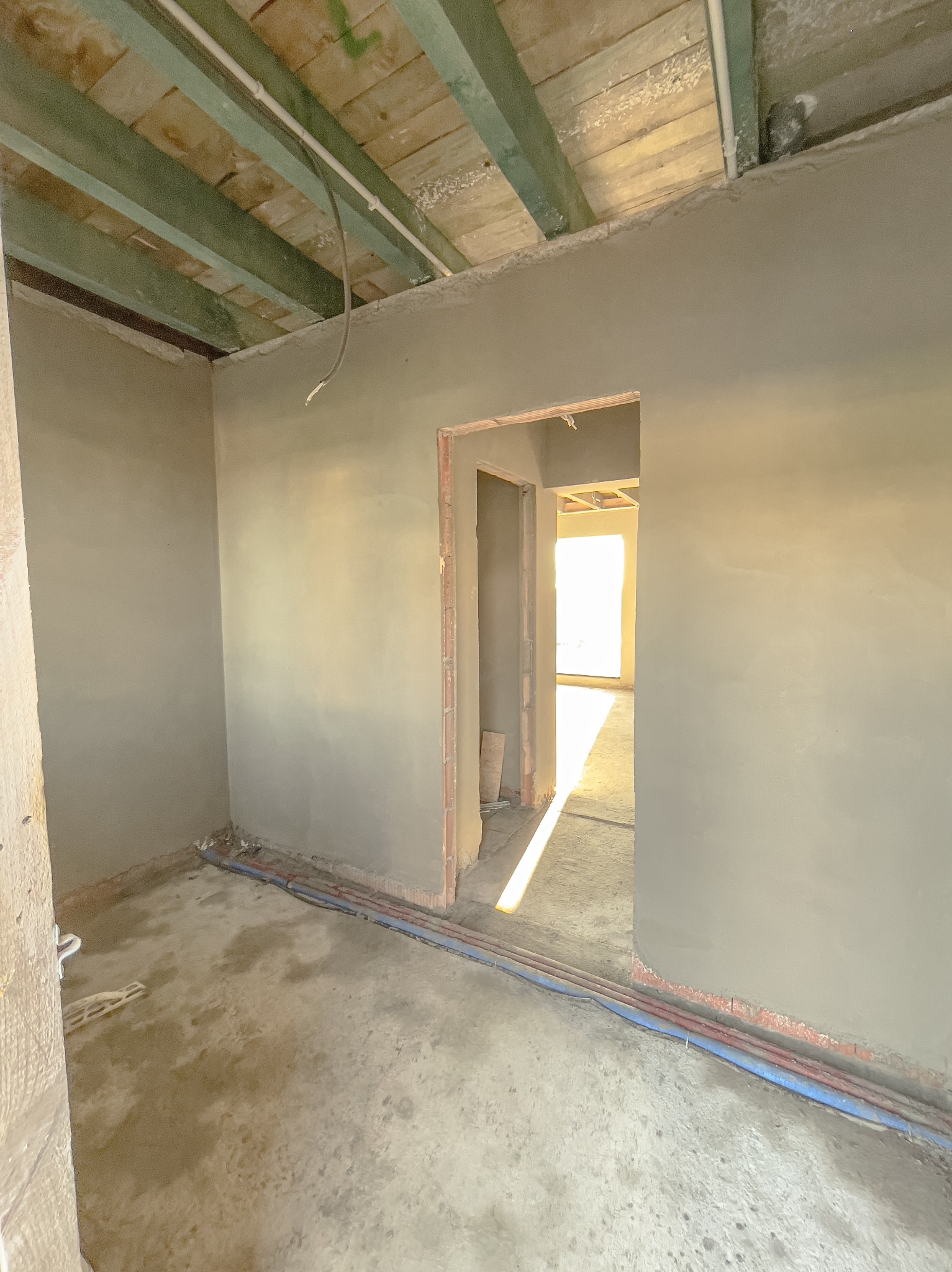 Wnętrze w stanie surowym po nałożeniu tynków; widoczne ściany pokryte szarym tynkiem, surowy strop z drewnianych belek, instalacje na podłodze i przejście do kolejnego pomieszczenia.