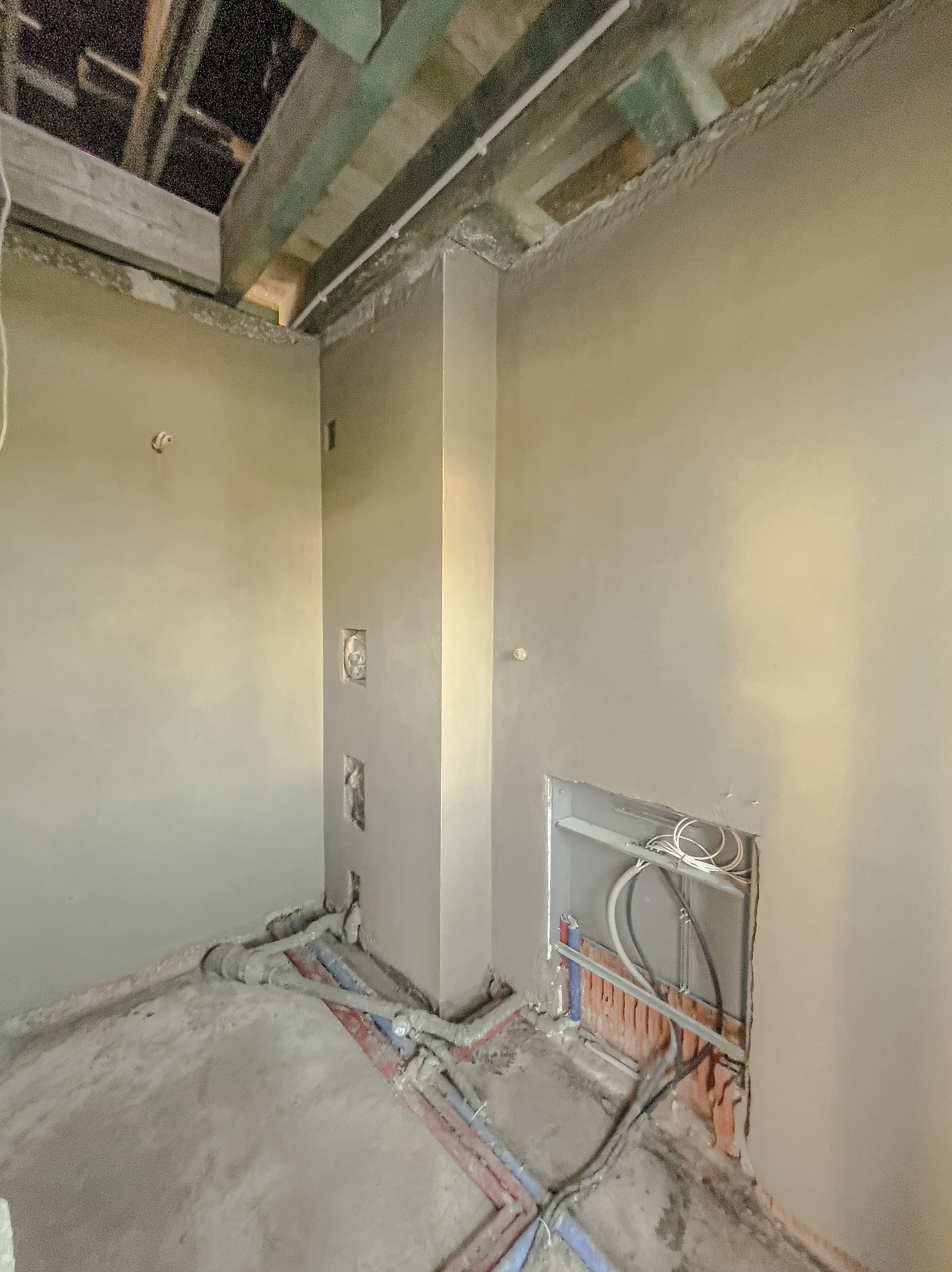 Wnętrze w trakcie tynkowania maszynowego: świeżo nałożony tynk na ścianach, widoczne instalacje elektryczne i hydrauliczne w bruzdach, surowy strop z drewnianych belek.