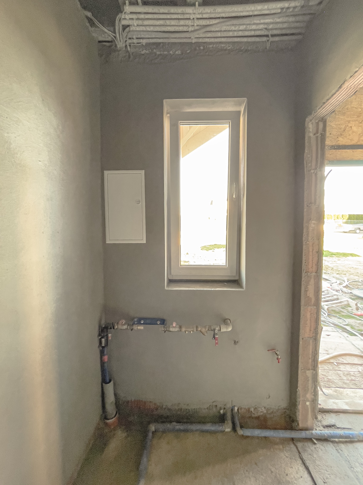 Świeżo nałożony, jasnoszary tynk maszynowy na ścianie wewnętrznej, widoczne instalacje wodne i elektryczne, okno z widokiem na zewnątrz, surowy stan wykończenia.