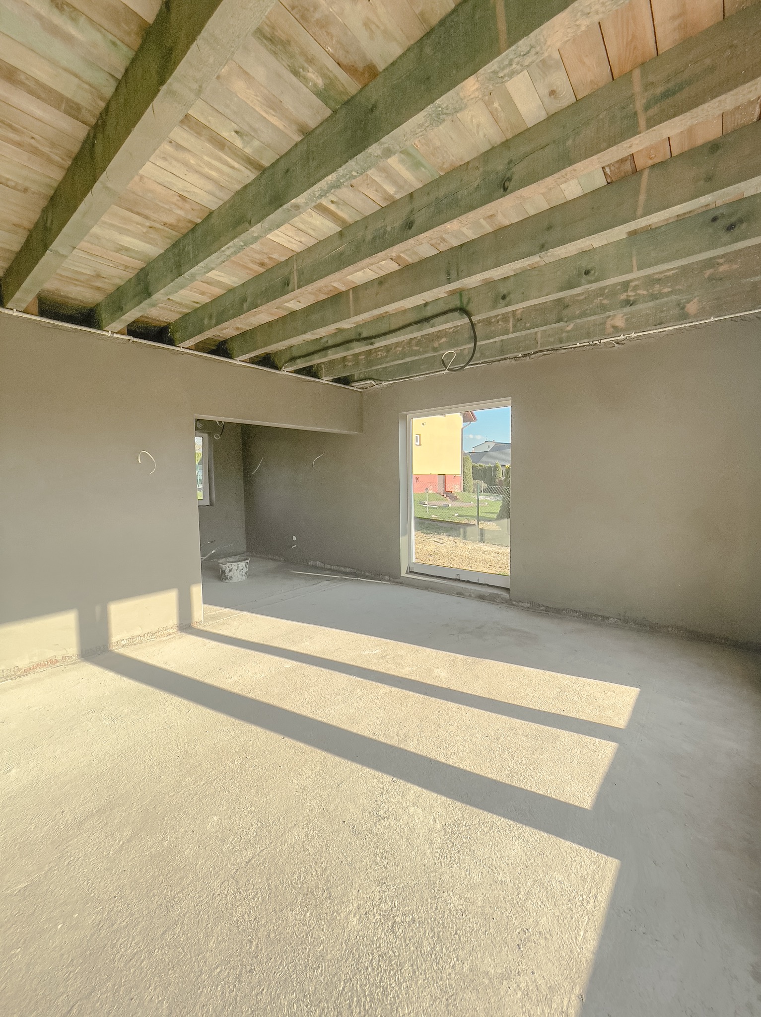 Wnętrze surowego domu po nałożeniu tynków, z widocznymi drewnianymi belkami stropowymi i promieniami słońca wpadającymi przez otwór drzwiowy.