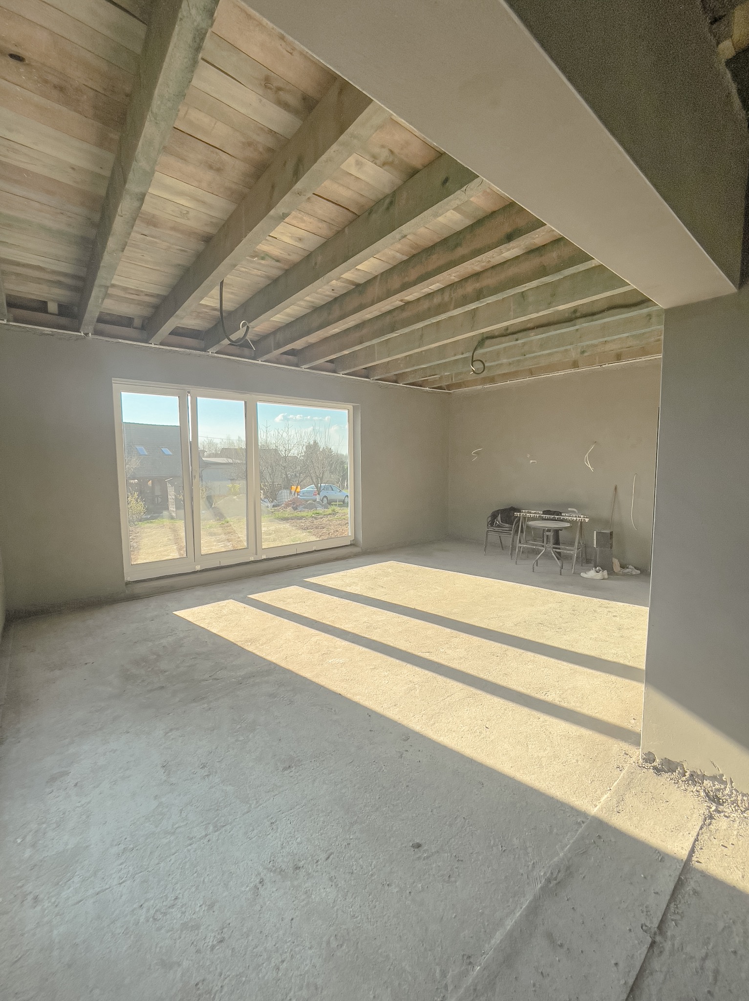 Surowe wnętrze domu w stanie deweloperskim po nałożeniu tynków, z widoczną drewnianą konstrukcją stropu i promieniami słońca wpadającymi przez duże okno tarasowe.