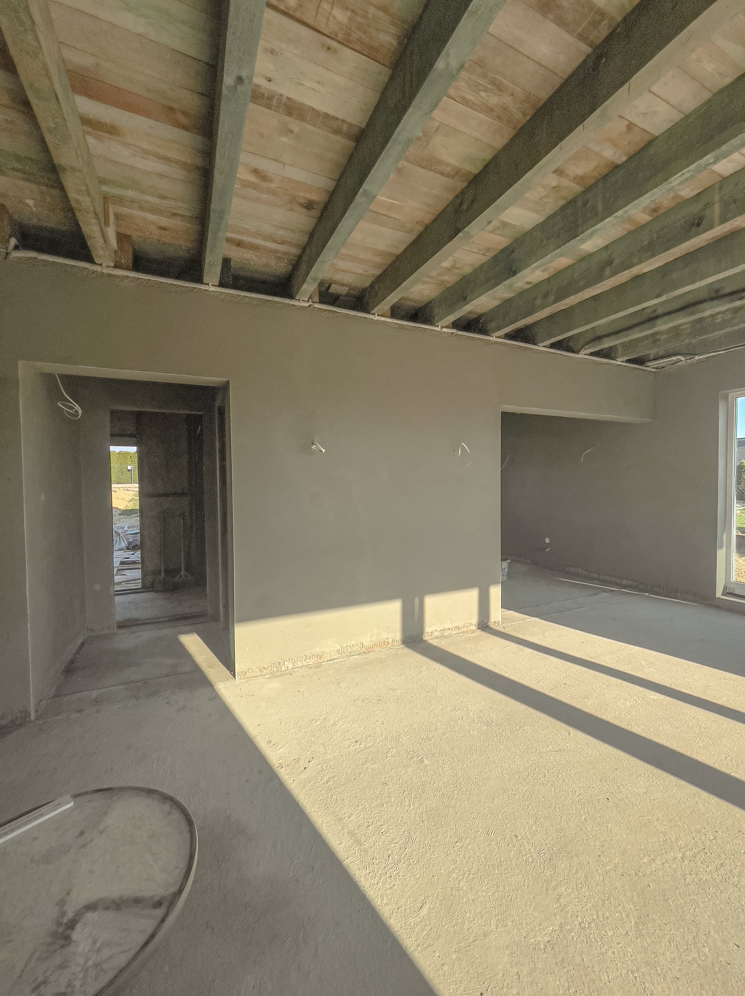 Surowe wnętrze domu w trakcie prac wykończeniowych, świeżo otynkowane ściany pod drewnianym stropem, widoczne kable i wpadające promienie słoneczne.