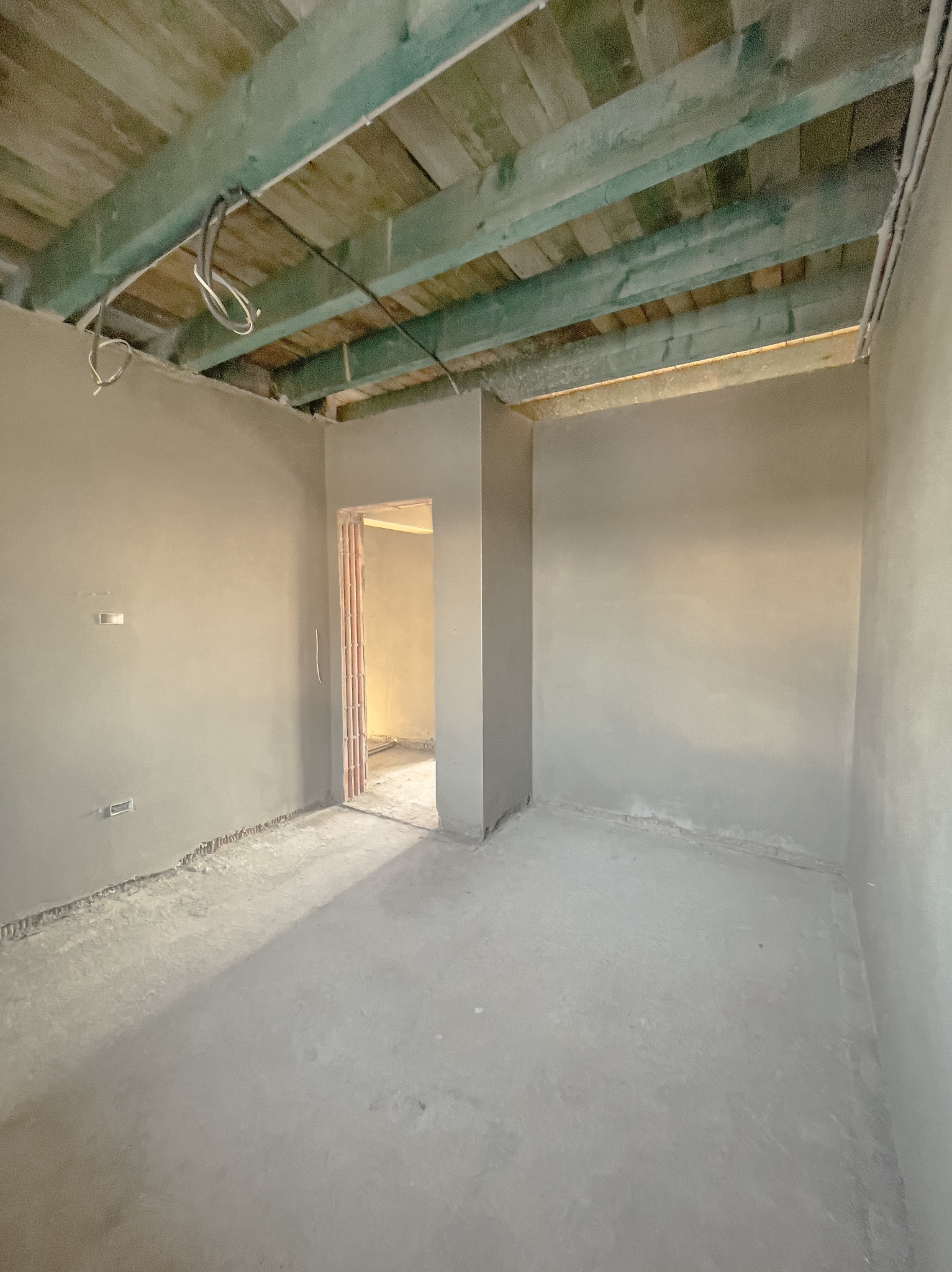 Wnętrze pokoju po nałożeniu szarego tynku maszynowego na ściany i podłogę, widoczne surowe drewniane belki stropowe pomalowane na zielono oraz wystające przewody elektryczne.