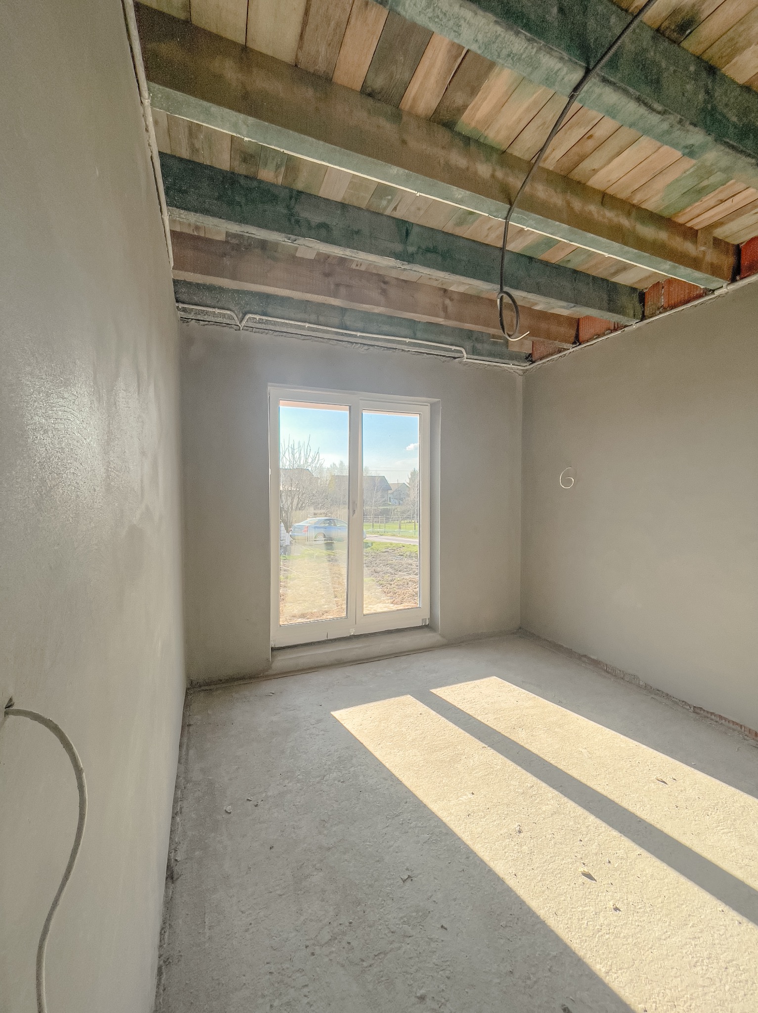 Wnętrze surowego pomieszczenia po nałożeniu tynków, widoczne drewniane elementy stropu, okno balkonowe z widokiem na podwórze, promienie słoneczne padające na podłogę.