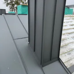 Szary dach z blachy trapezowej z kominem w kolorze antracytu, widoczne deski podkładowe pokryte śniegiem, w tle drzewo z jemiołą.