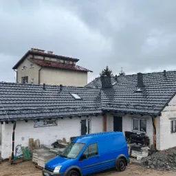 Dom w trakcie budowy z nowym, ciemnoszarym dachem z dachówki ceramicznej, widoczny niebieski samochód dostawczy, na ziemi leżą materiały budowlane.