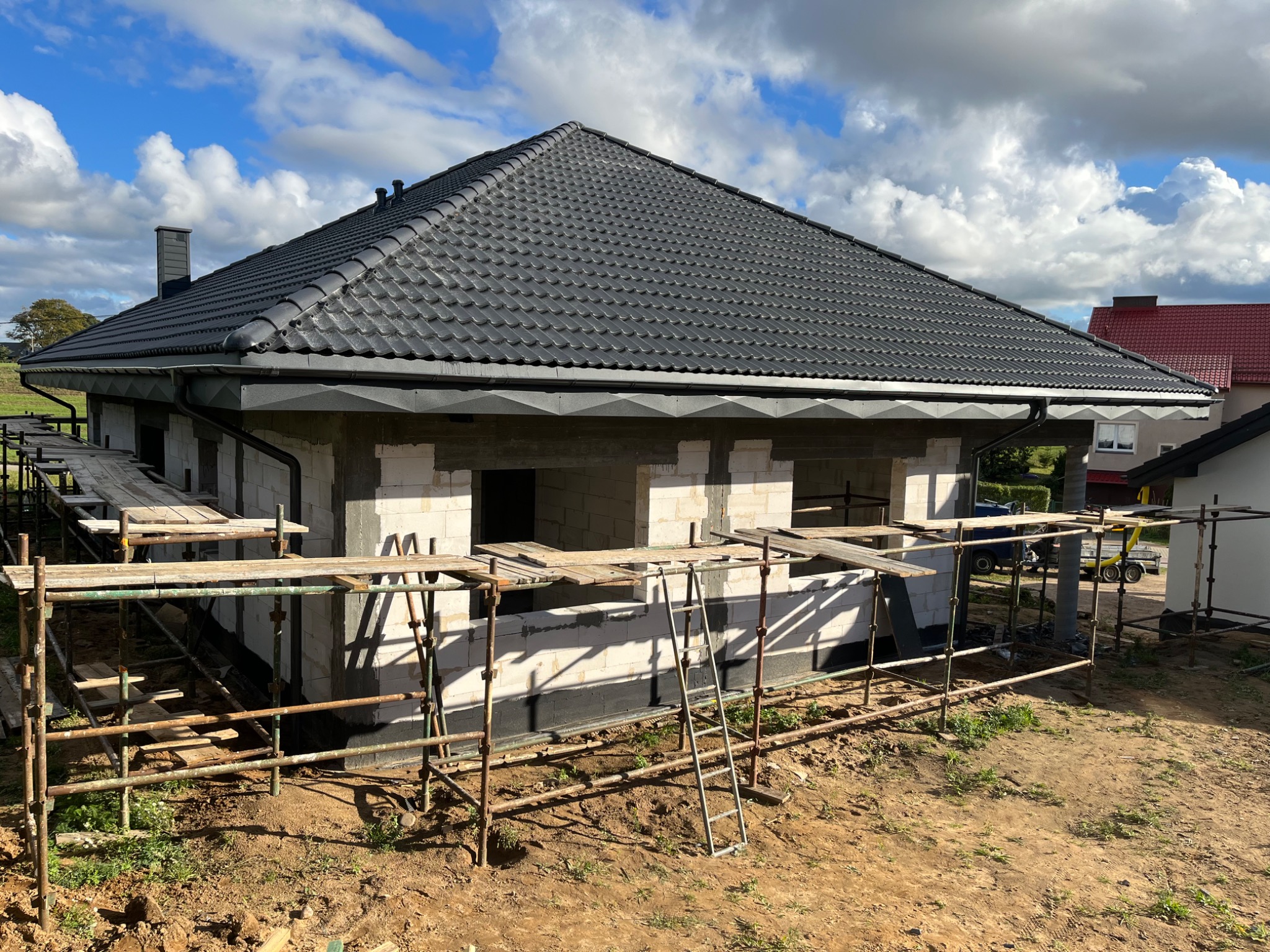 Nowy, grafitowy dach z dachówki ceramicznej na surowym budynku w trakcie budowy, widoczne rusztowania i elementy wykończeniowe.