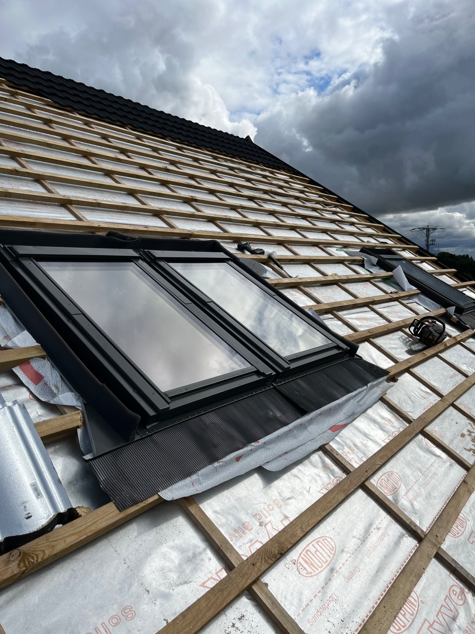 Dach w trakcie budowy z zamontowanymi oknami dachowymi, widoczne drewniane łaty, membrana dachowa i częściowo ułożone czarne dachówki. W tle pochmurne niebo.