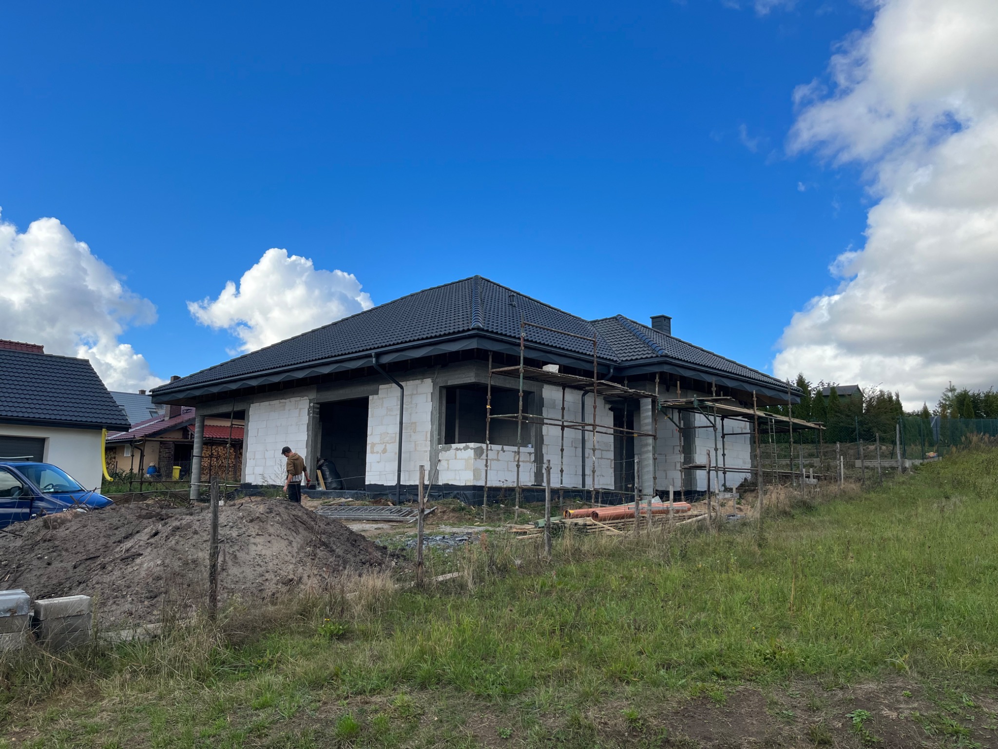 Dom w trakcie budowy z czarnym dachem z dachówki ceramicznej, widoczne rusztowania i stos materiałów budowlanych na działce, błękitne niebo z chmurami w tle.