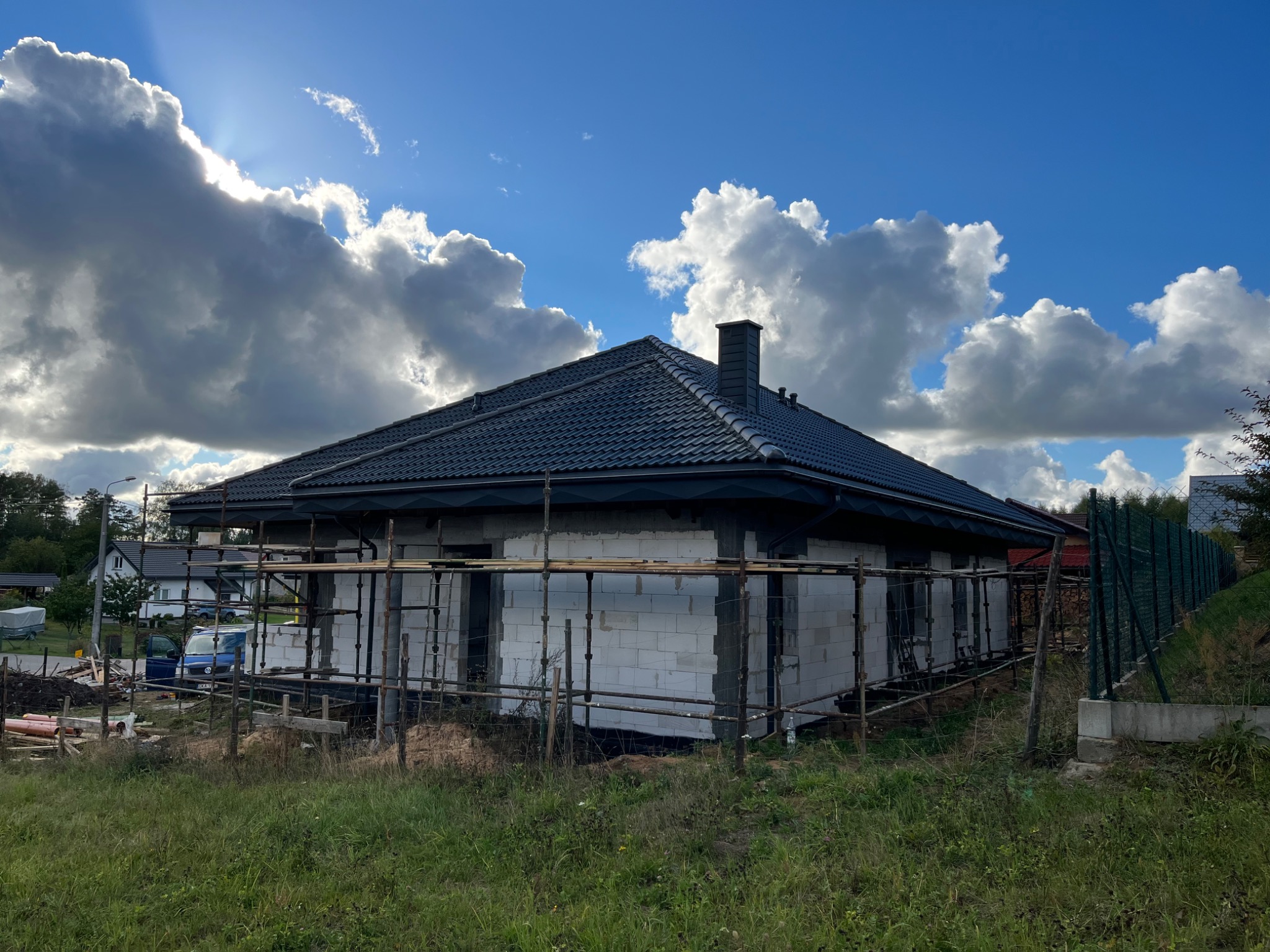 Dom jednorodzinny w trakcie budowy, z widocznym szarym dachem z dachówki ceramicznej i kominem, otoczony rusztowaniem, na tle błękitnego nieba z malowniczymi chmurami.