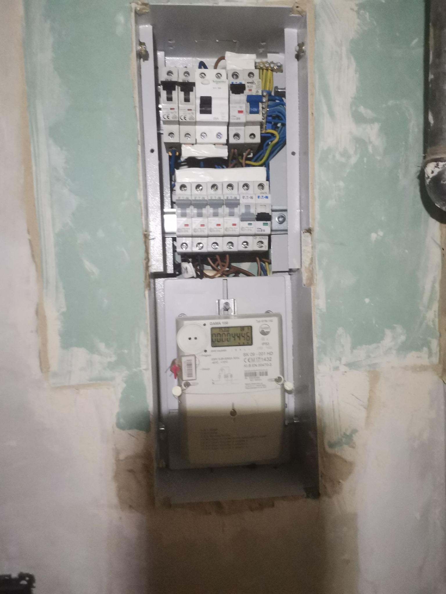 Szara skrzynka z bezpiecznikami i licznikiem energii elektrycznej zamontowana w surowej wnęce ściany z widocznymi śladami gładzi szpachlowej.