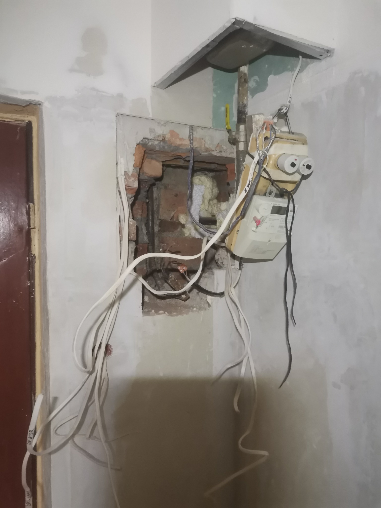 Niedokończona instalacja licznika elektrycznego w ścianie z widocznymi przewodami i fragmentami muru, w trakcie prac remontowych.
