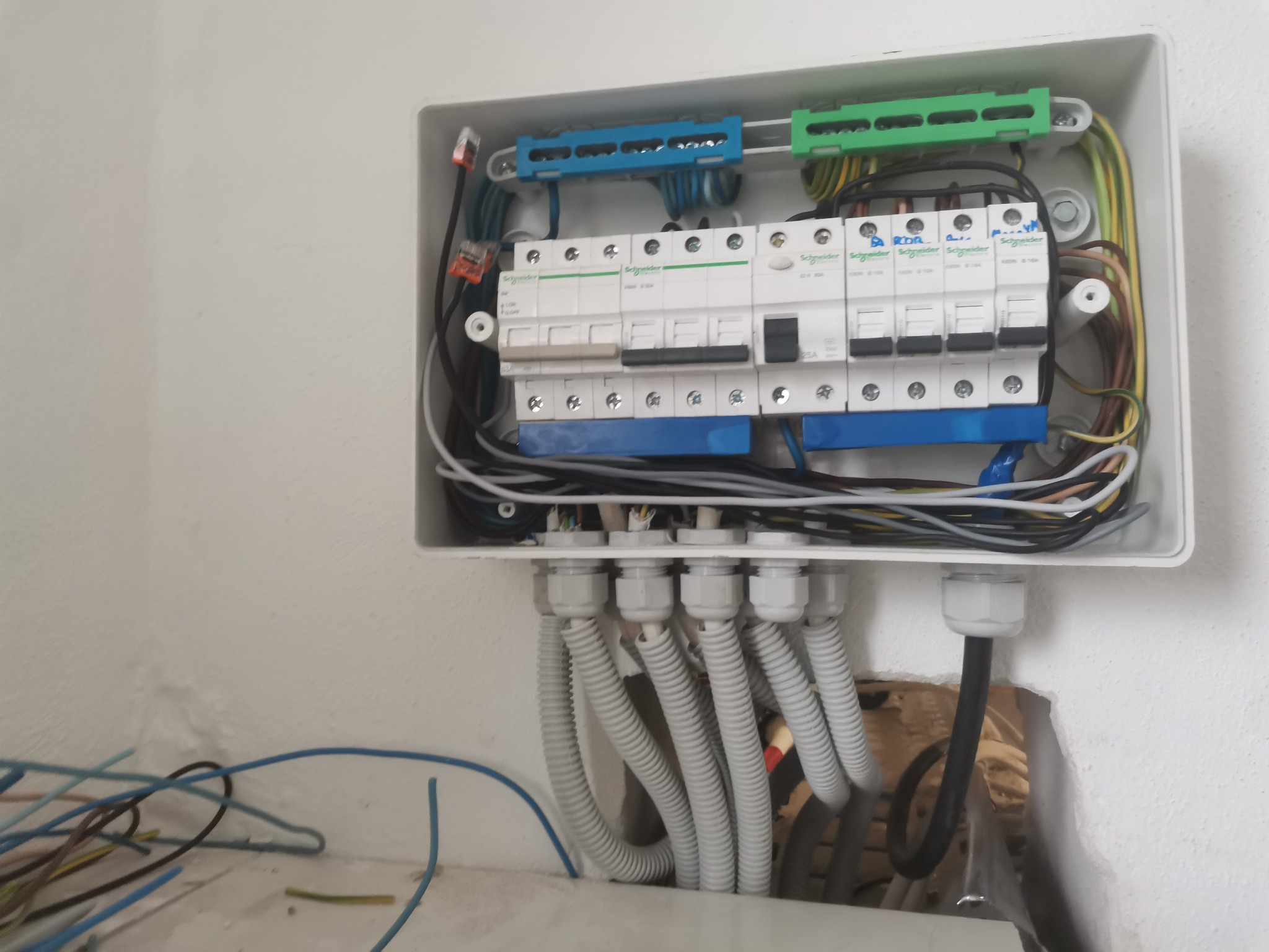 Otwarta skrzynka bezpiecznikowa Schneider z widocznymi bezpiecznikami, przewodami w różnych kolorach i peszlami, zamontowana na białej ścianie, z luźno leżącymi przewodami na podłodze.