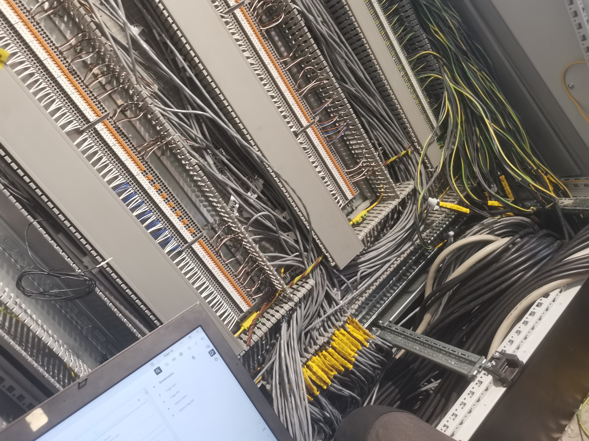 Rozbudowana szafa sterownicza w trakcie montażu, widoczne liczne kable szare, czarne i żółto-zielone, listwy zaciskowe oraz laptop z otwartą stroną internetową w lewym dolnym rogu.