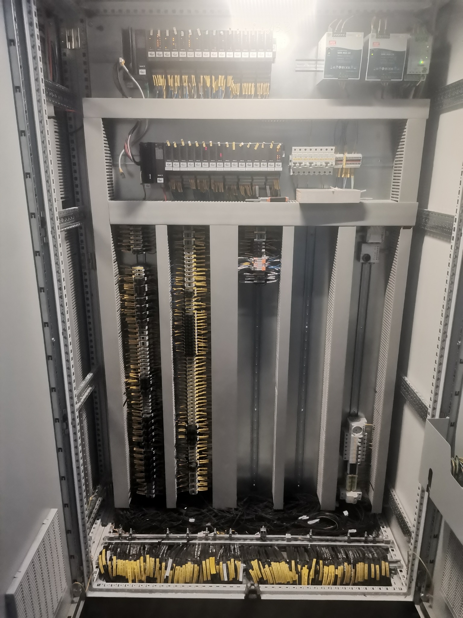 Wnętrze szafy sterowniczej z licznymi przewodami oznaczonymi żółtymi etykietami, widoczne moduły sterujące i zasilacze, uporządkowane okablowanie w szynach montażowych.