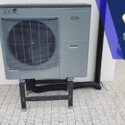 Zewnętrzna jednostka pompy ciepła NIBE AMS 10-12 zamontowana na metalowej konstrukcji przed białą ścianą budynku w Kościerzynie, widoczny fragment trawnika i kostki brukowej.