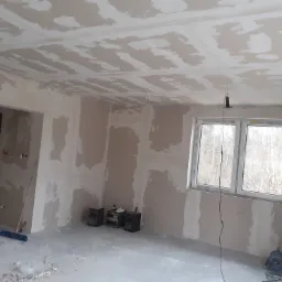 Pok&oacute;j w trakcie prac wykończeniowych: ściany i sufit pokryte wstępną warstwą gładzi, widoczne narzędzia, sprzęt audio pod ścianą, okno z widokiem na drzewa.