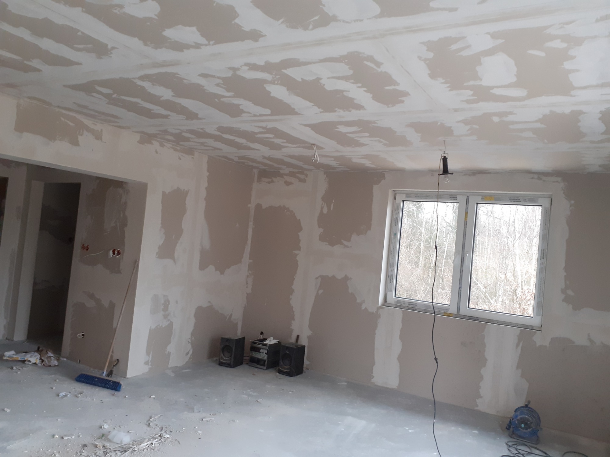 Pokój w trakcie prac wykończeniowych: ściany i sufit pokryte wstępną warstwą gładzi, widoczne narzędzia, sprzęt audio pod ścianą, okno z widokiem na drzewa.