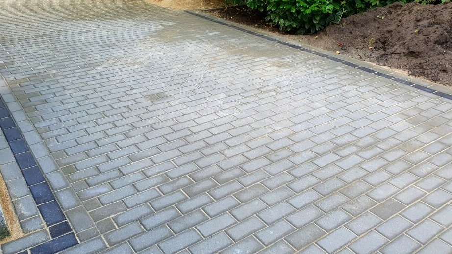 Ułożona, szara kostka brukowa w jodełkę z ciemnoszarym obramowaniem, widoczna krawędź trawnika z zielenią i kopiec ziemi w tle.