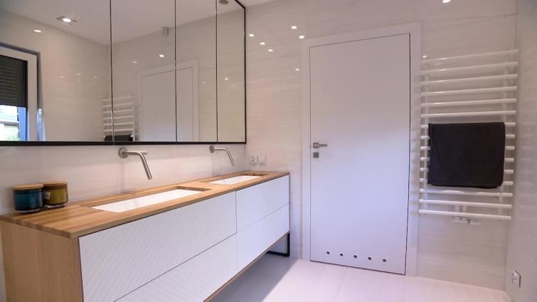 Nowoczesna łazienka z podwójną umywalką na drewnianym blacie, minimalistycznym lustrem z czarną ramą, białymi szafkami z ukośnymi frezowaniami, grzejnikiem drabinkowym z szarym ręcznikiem i białymi...