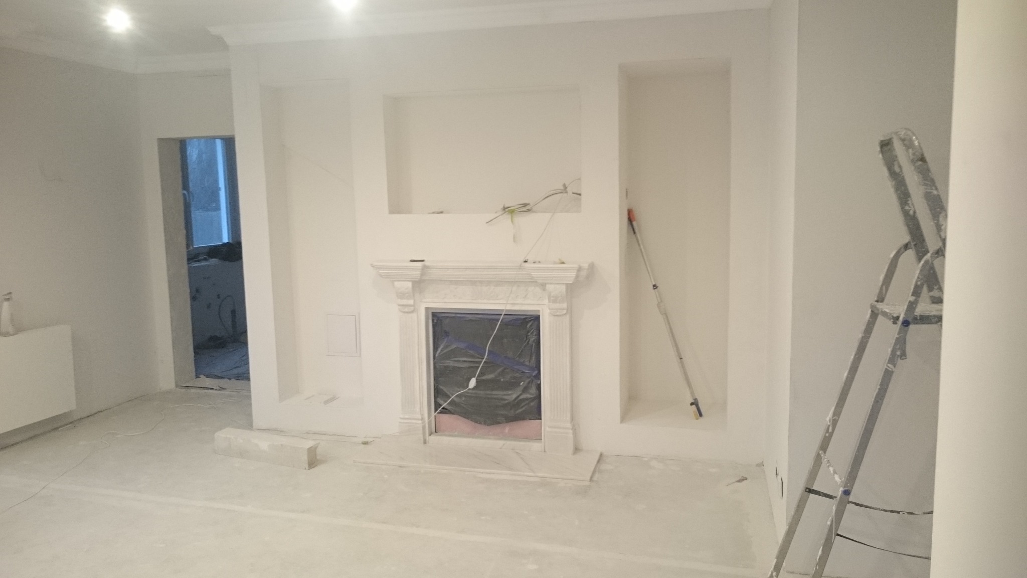 Wnętrze w trakcie malowania na biało, widoczny kominek zabezpieczony folią, drabina malarska i wałek do malowania, podłoga pokryta resztkami farby.