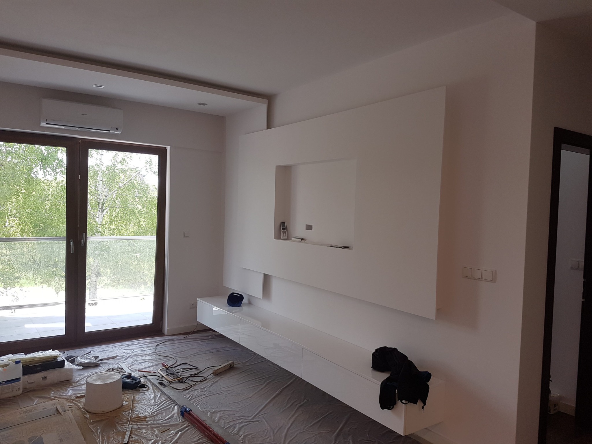 Nowoczesny salon w trakcie wykańczania: białe ściany, zabudowa TV z półkami, zabezpieczona folią podłoga i narzędzia pracy.