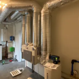 Wnętrze kotłowni z zainstalowanym systemem wentylacji z rurami spiro, kotłem grzewczym i zasobnikiem ciepłej wody, widoczne instalacje wodne i gazowe.