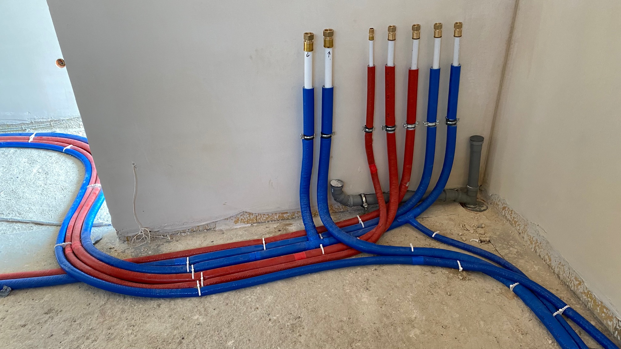 Nowa instalacja wodna z rur PEX w kolorach czerwonym i niebieskim, prowadząca wzdłuż ściany i podłogi w surowym stanie, z widocznymi mocowaniami i przyłączami.
