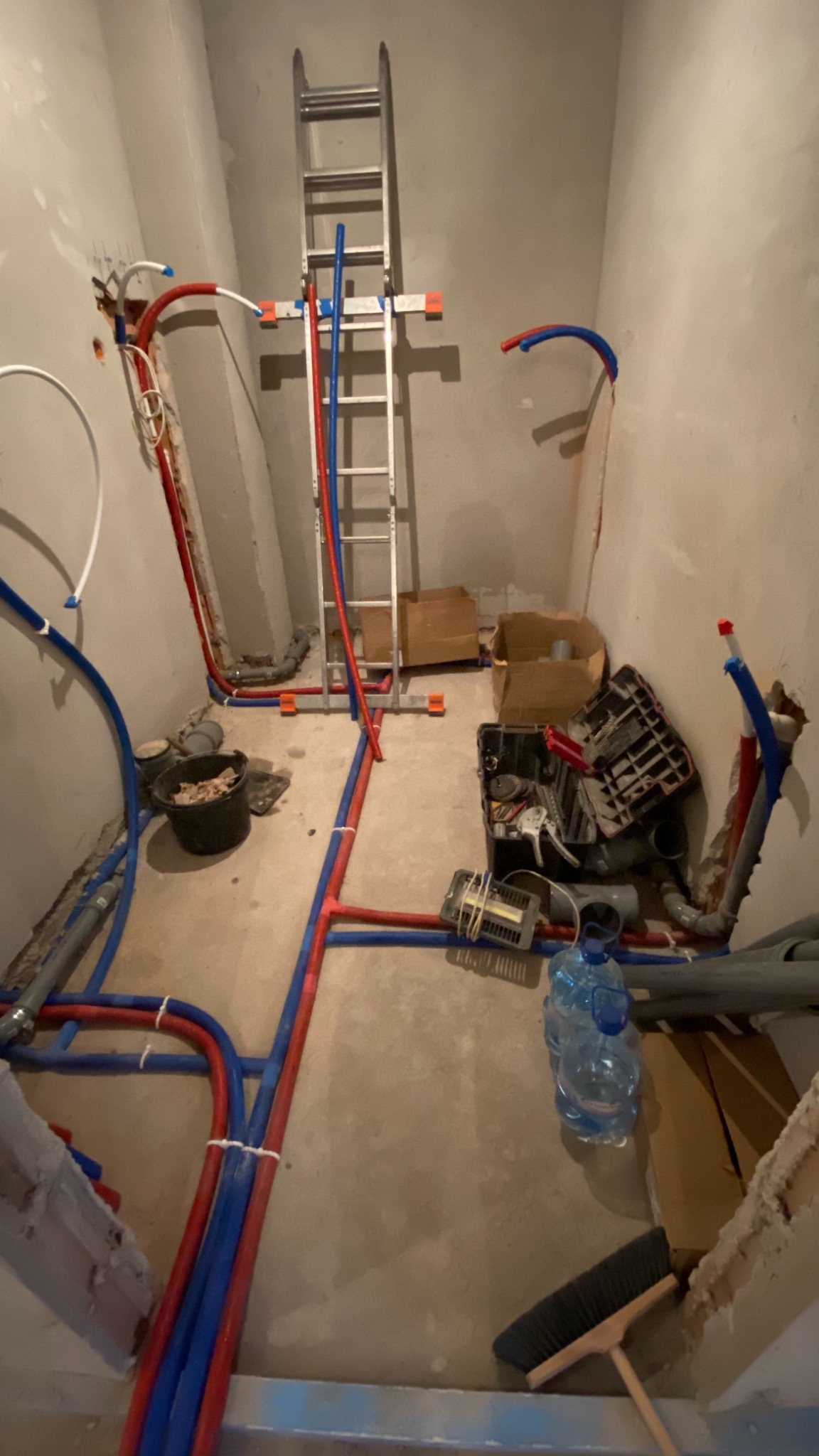 Wnętrze w trakcie prac hydraulicznych: widoczne rury PEX w kolorach czerwonym i niebieskim prowadzone po ścianach i podłodze, drabina aluminiowa oparta o ścianę, wiadro z odpadami, skrzynka...