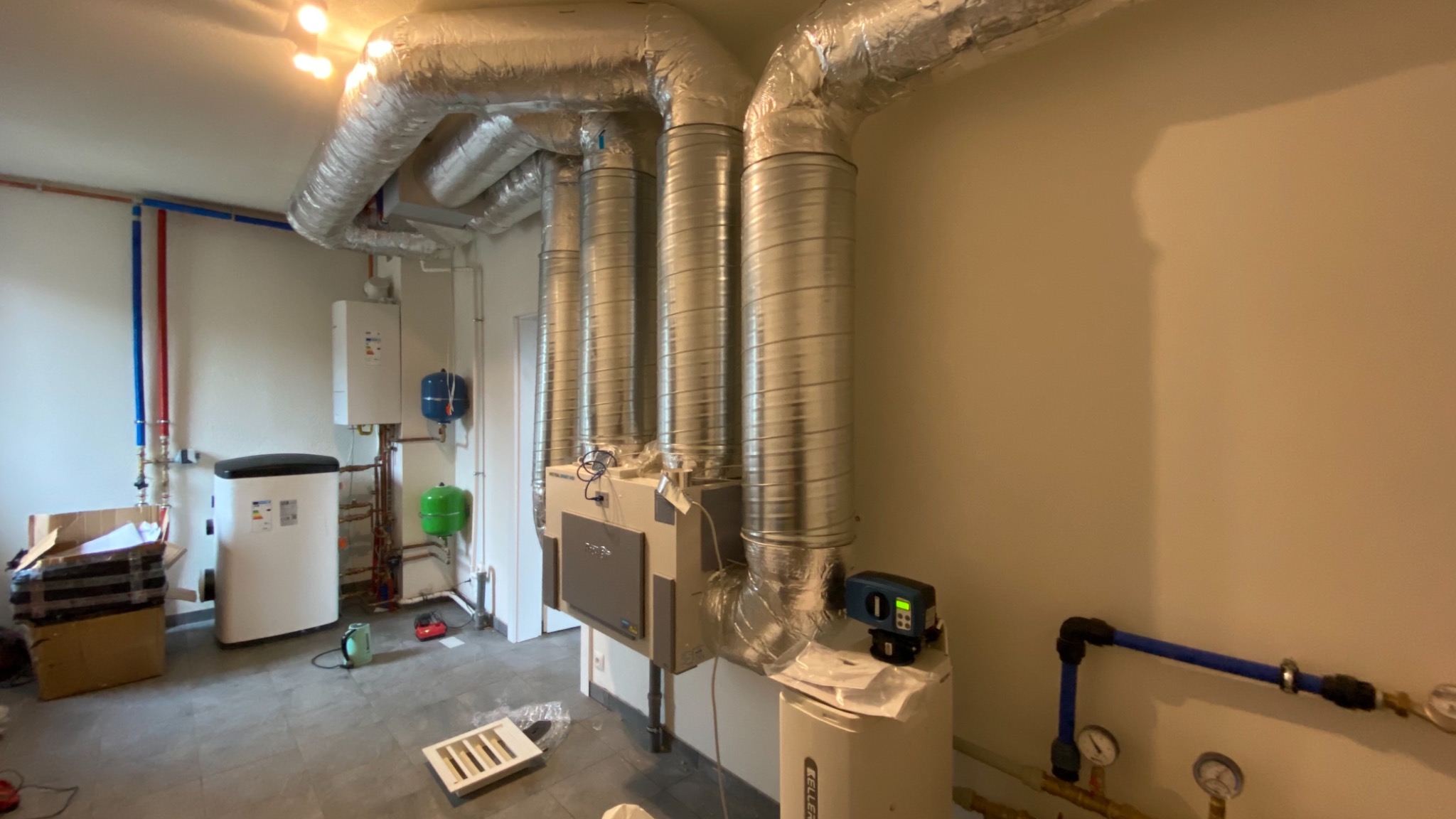 Wnętrze kotłowni z zainstalowanym systemem wentylacji z rurami spiro, kotłem grzewczym i zasobnikiem ciepłej wody, widoczne instalacje wodne i gazowe.