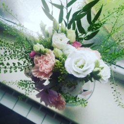 Amelia Pracownia Florystyczna - Kompozycja kwiatowa z białych i różowych róż oraz zielonych dodatków w szklanym wazonie, ustawiona na parapecie okiennym z widocznymi płytkami.