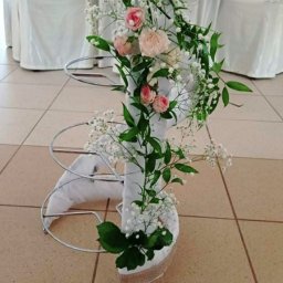 Amelia Pracownia Florystyczna - Dekoracja kwiatowa z różowych i białych róż, gipsówki i zielonych liści, umieszczona na metalowym stelażu owiniętym białą tkaniną, stojąca na kafelkowanej podłodze.