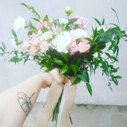 Amelia Pracownia Florystyczna - Delikatny bukiet ślubny z różowych róż, białych goździków i zielonych liści, przewiązany jutową wstążką, trzymany w dłoni z tatuażem kwiatu.