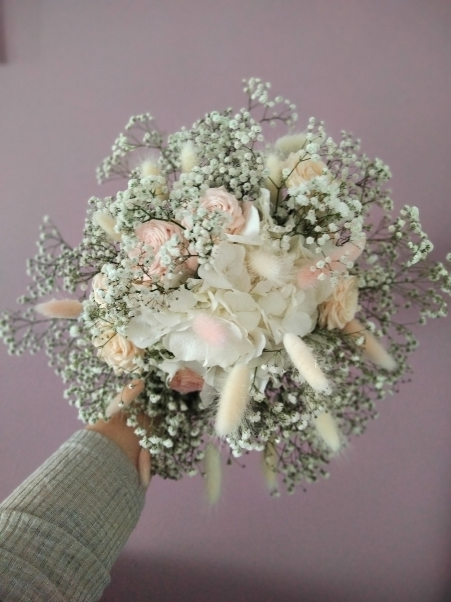 Delikatny bukiet ślubny z białych i różowych kwiatów, gipsówki i suszonych traw, trzymany w dłoni na tle fioletowej ściany.