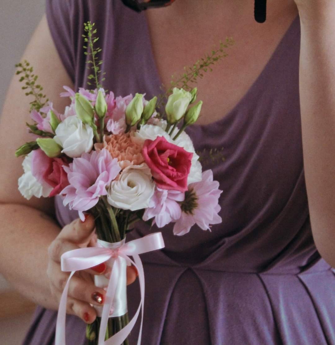 Kobieta w fioletowej sukience trzyma delikatny bukiet ślubny z różowych i białych kwiatów, przewiązany jasnoróżową wstążką, przygotowany w Legionowie.