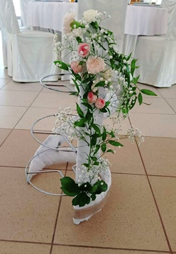 Dekoracja kwiatowa z różowych i białych róż, gipsówki i zielonych liści, umieszczona na metalowym stelażu owiniętym białą tkaniną, stojąca na kafelkowanej podłodze.