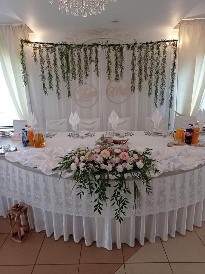 Uroczysty stół weselny z białym obrusem, kwiatową kompozycją z różowych i białych róż, dekoracją z zielonych girland i drewnianymi imionami pary młodej na białym tle.