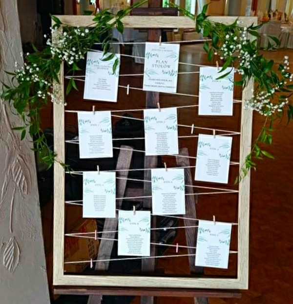 Plan stołów weselnych, karty z nazwiskami gości przypięte klamerkami do sznurków rozpiętych w drewnianej ramie, dekoracja z białych kwiatów i zielonych liści