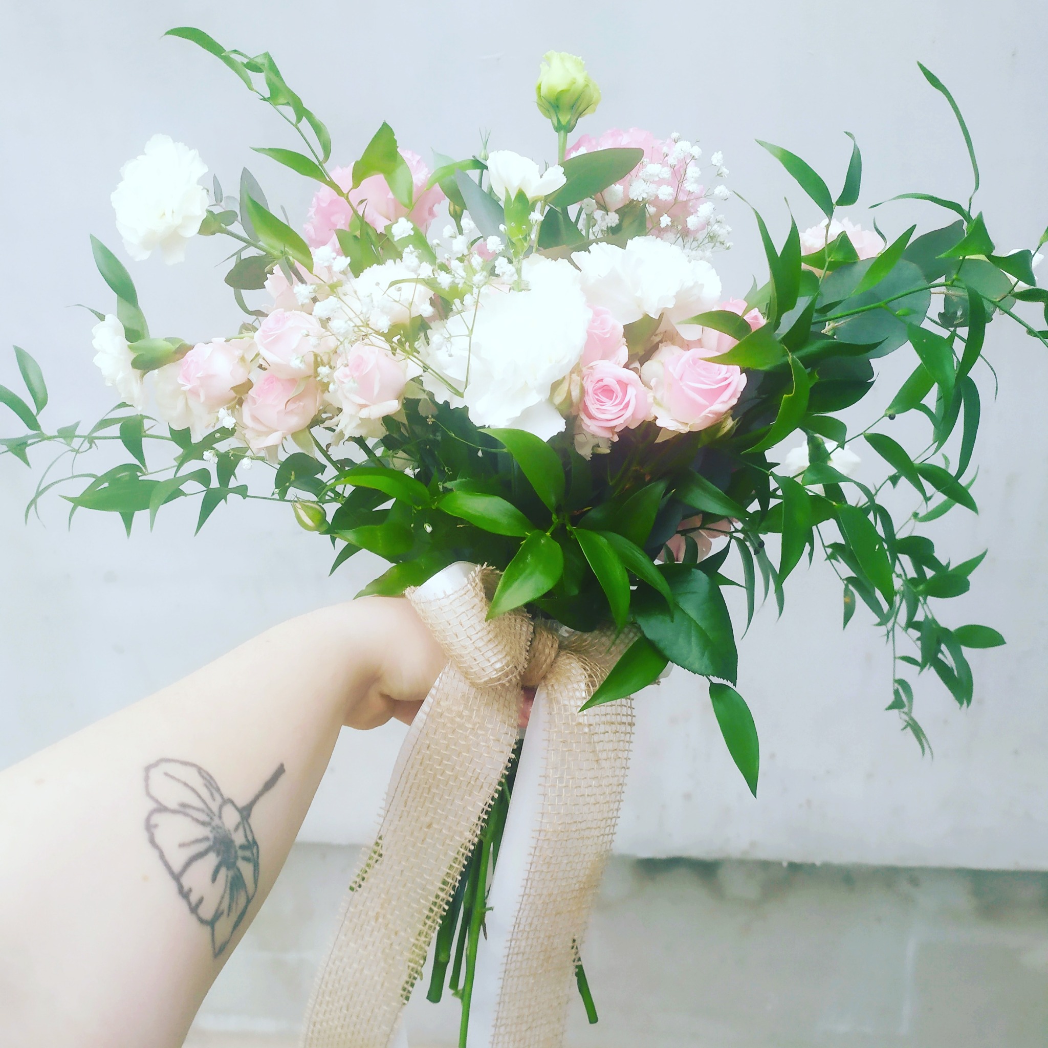 Delikatny bukiet ślubny z różowych róż, białych goździków i zielonych liści, przewiązany jutową wstążką, trzymany w dłoni z tatuażem kwiatu.