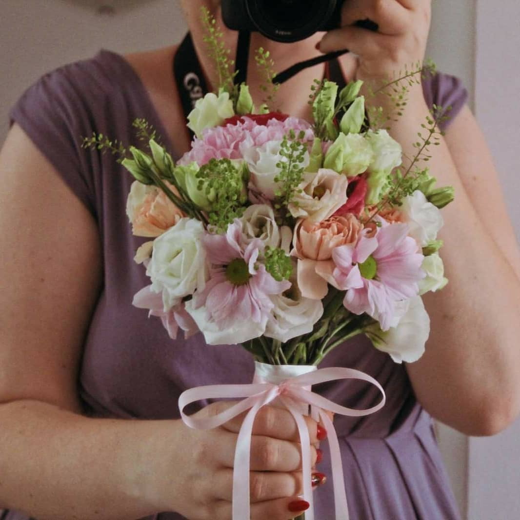Osoba w fioletowej sukience trzyma bukiet z różowych, białych i brzoskwiniowych kwiatów, robiąc zdjęcie aparatem Canon.