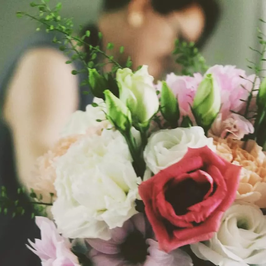 Delikatny bukiet kwiatów w pastelowych odcieniach różu, bieli i kremu z pojedynczą czerwoną różą na pierwszym planie, na rozmytym tle sylwetka osoby.