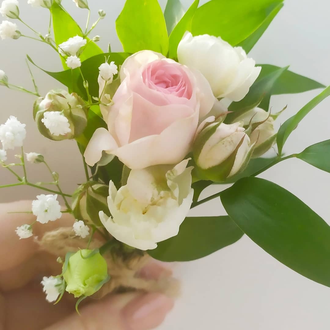 Delikatny butonierka z różowej i białych róż, gipsówki i zielonych liści, owinięta sznurkiem jutowym, trzymana w dłoni na jasnym tle.