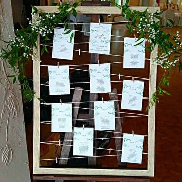 Rustykalny plan stołów weselnych w drewnianej ramie, ozdobiony zielenią i białymi kwiatami, z kartkami przypiętymi klamerkami.