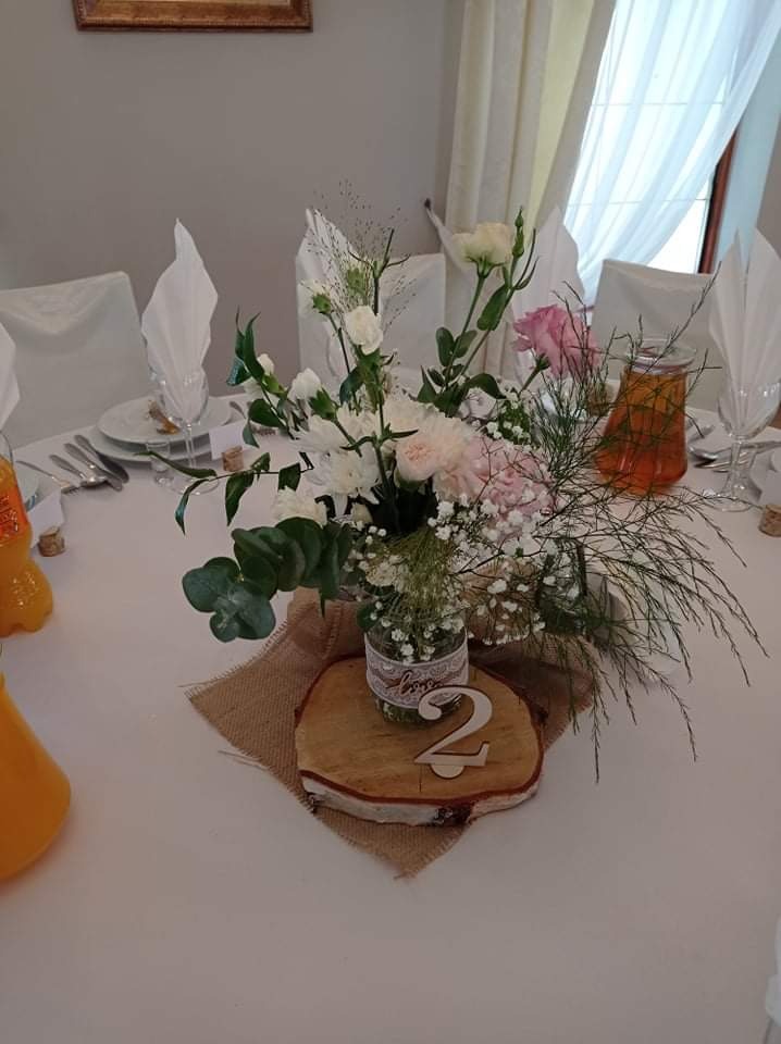 Dekoracja stołu weselnego: kompozycja kwiatowa w słoiku z koronką, numer stołu '2' na drewnianym plastrze, jutowy bieżnik, zastawa i napoje w tle.